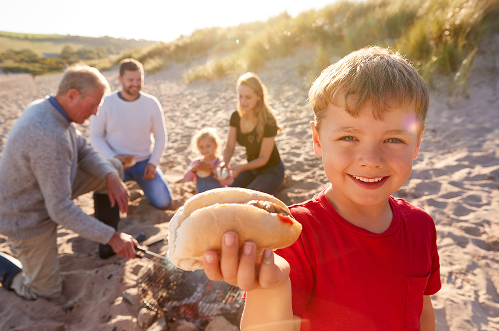 A young boy in red t-shirt smiling with a burger in his hand on a beach with sand dunes behind and family sitting on blanket