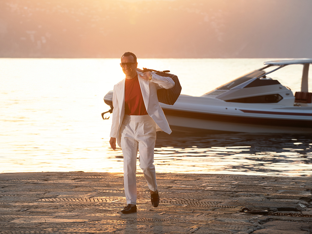 Alessandro Martorana walks waterside at sunset in white suit, with GranTurismo 12.5 AMare moored behind.