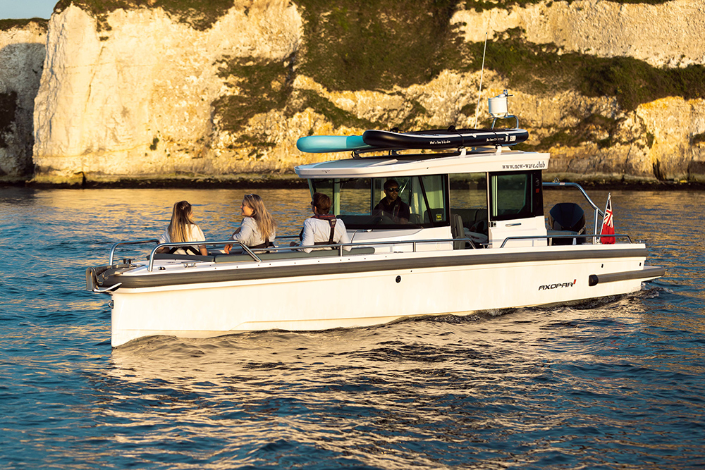 Axopar boat cruising along sunlit chalk cliffs with passengers seated on deck and paddleboards stowed on the roof.