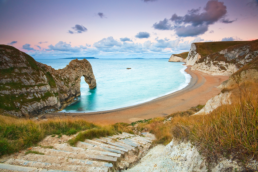 Durdle Door’s natural rock arch and curved beach at sunset, viewed from the cliff path overlooking turquoise water.