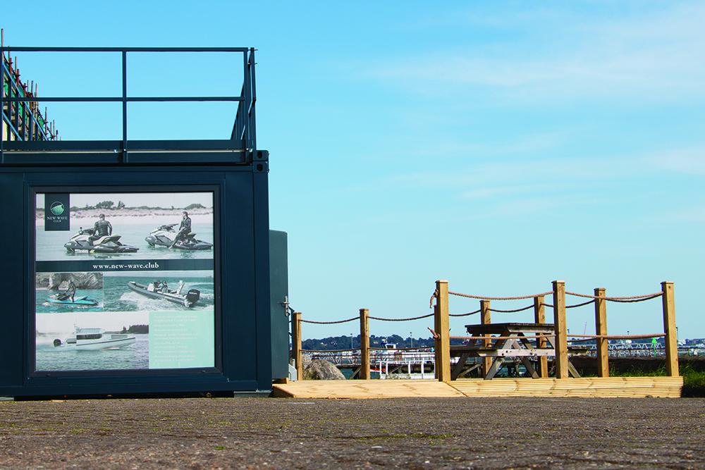 New Wave Club’s waterside facility with decking and picnic seating overlooking the marina under a clear blue sky.
