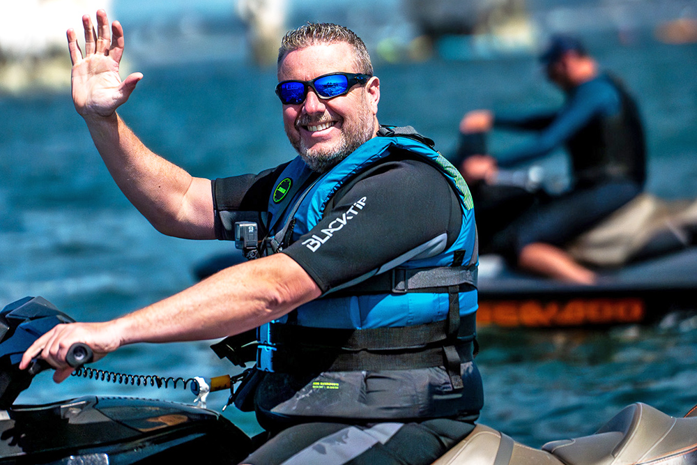 Smiling man in wetsuit and lifejacket waves while riding a jet ski on calm water, with another rider in the background.